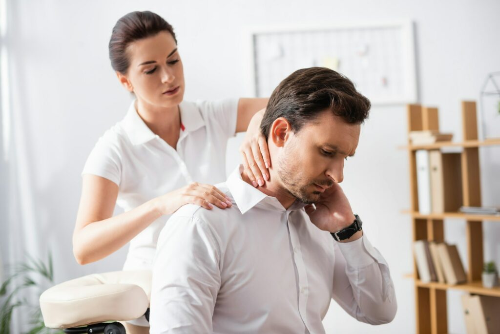 Female massage therapist massaging hurting neck of businessman sitting on massage chair on blurred